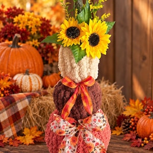May include: A stacked pumpkin decoration made of three textured, crocheted spheres in cream, brown, and rust colors. The top sphere is topped with two yellow sunflowers and green foliage. An orange plaid ribbon and a floral print ribbon are tied around.