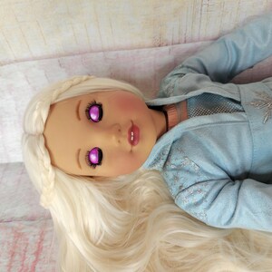 May include: A doll with long, white, braided hair and a light blue, sparkly dress. The doll has pink cheeks and closed eyes with purple irises.
