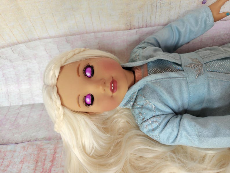 May include: A doll with long, white, braided hair and a light blue, sparkly dress. The doll has pink cheeks and closed eyes with purple irises.