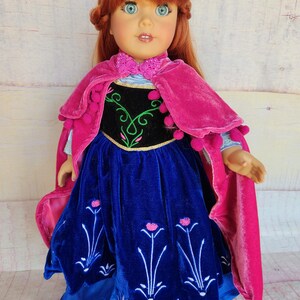 May include: A red-haired doll wearing a blue velvet dress with pink flowers embroidered on the skirt. The doll is also wearing a pink velvet cape with pom-poms around the edges.