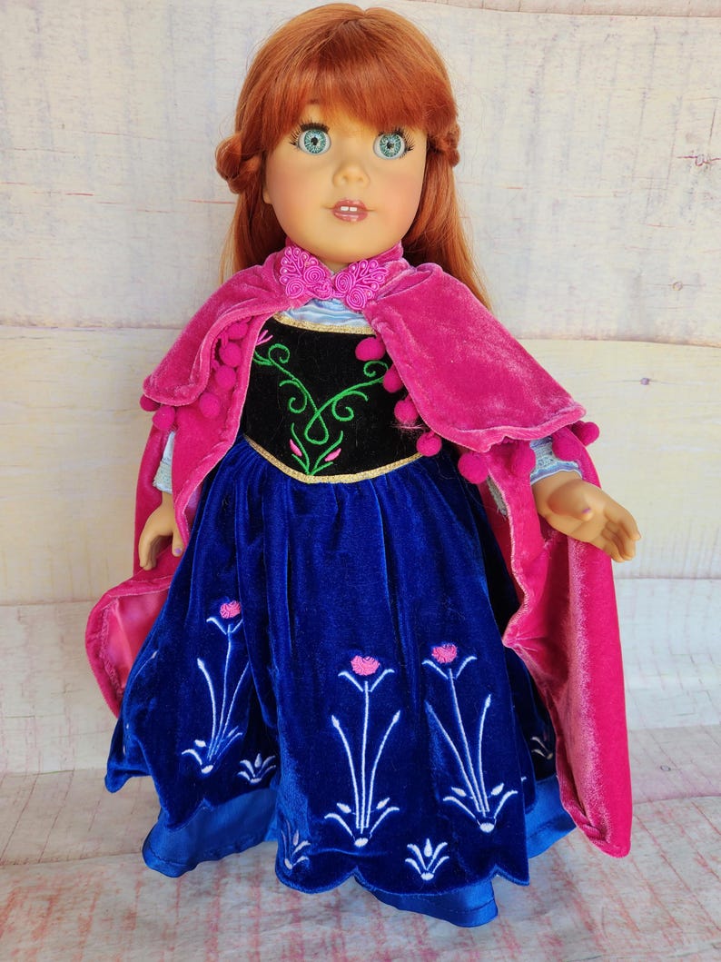 May include: A red-haired doll wearing a blue velvet dress with pink flowers embroidered on the skirt. The doll is also wearing a pink velvet cape with pom-poms around the edges.