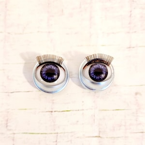 May include: Two silver-colored doll eyes with purple irises and black eyelashes. The eyes are set in a circular frame.