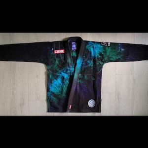 May include: A black and blue tie-dye Jiu-Jitsu gi with the text "CRNR" on the left sleeve and "CONQUER" on the right sleeve.