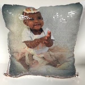 May include: A reversible sequin pillow featuring a photograph of a smiling baby. The sequins transition from silver to a gradient of silver and rose gold. The baby is wearing a white onesie and a floral headband.