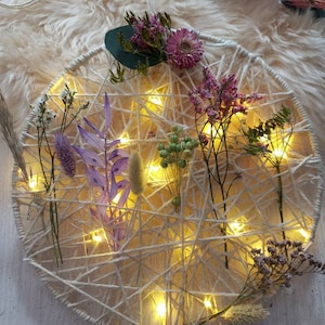 May include: A circular macrame wall hanging with dried flowers and string lights. The macrame is made with white string and the flowers are in shades of pink and purple.