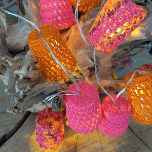 Puede incluir: Una cadena de luces con pantallas de lámparas de crochet en rosa, naranja y una mezcla de rosa y naranja. Las luces están enhebradas en una rama y están encendidas, proyectando un brillo cálido.
