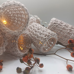 Könnte beinhalten: Eine Lichterkette mit gehäkelten Bezügen in beige. Die Lichter sind an und strahlen ein warmes Licht. Die Lichter befinden sich auf einem Zweig mit kleinen orangen Beeren.