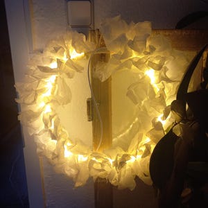 Könnte beinhalten: Ein handgefertigter, beleuchteter Kranz mit kreisförmigem Design. Der Kranz ist mit weißem, gerüschtem Papier und warmen, gelben LED-Lichtern verziert. Er ist auf einem Holzrahmen montiert und erzeugt ein weiches, einladendes Leuchten.