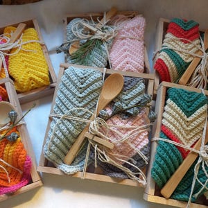 Könnte beinhalten: Holzkisten gefüllt mit farbenfrohen, gehäkelten Waschlappen und Holzlöffeln. Jeder Waschlappen hat eine andere Farbe, darunter Gelb, Orange, Rosa, Grün und Grau. Die Waschlappen sind mit Bindfäden gebunden und mit Zweigen verziert.