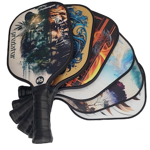 Könnte beinhalten: Eine Sammlung von Pickleball-Schlägern mit schwarzen Griffen und verschiedenen künstlerischen Designs. Ein Schläger zeigt einen Löwen und einen Tiger, während andere abstrakte Kunst und einen Phönix zeigen. Das Wort "predator" ist auf einem Schläger aufgedruckt.