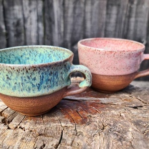 May include: Two handmade ceramic mugs with speckled glaze. One mug is a light green with brown speckles and the other is a light pink with brown speckles. Both mugs have a brown rim and a brown handle.