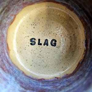 May include: Close-up of the inside of a ceramic mug with a light brown glaze and a dark brown rim. The word "SLAG" is printed in black on the bottom of the mug.