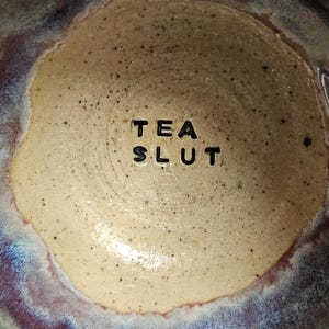 May include: A close-up of the inside of a ceramic teacup with a light brown glaze and a dark purple rim. The words "TEA SLUT" are written in black lettering on the bottom of the cup.