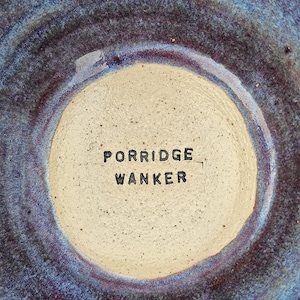 May include: A ceramic bowl with a speckled beige centre featuring the stamped black text PORRIDGE WANKER. The bowl has a textured, mottled glaze in shades of deep blue, purple, and indigo that radiates outwards from the central beige circle.