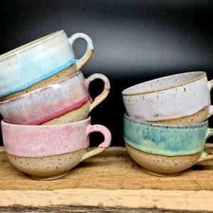 May include: Four ceramic mugs with speckled glaze in various shades of blue, pink, and green. The mugs are stacked on top of each other and have a rustic, handmade look.