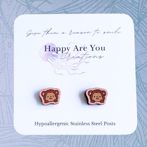 May include: A pair of wooden earrings featuring a cartoon monkey design. The earrings are pink and brown and have hypoallergenic stainless steel posts. The card reads "Give them a reason to smile. Happy Are You Creations. Hypoallergenic Stainless Steel Posts."