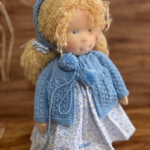 Puede incluir: Una muñeca hecha a mano con cabello rubio rizado, que lleva un cárdigan azul tejido y un vestido blanco con estampado floral. La muñeca tiene ojos azules y una expresión dulce.