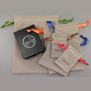 May include: An assortment of five beige velvet drawstring pouches with different coloured ribbons. The pouches are arranged in decreasing size, with the largest pouch at the back and the smallest pouch at the front. The pouches are all labelled with the text "LIALUCINE FINE JEWELLERY".