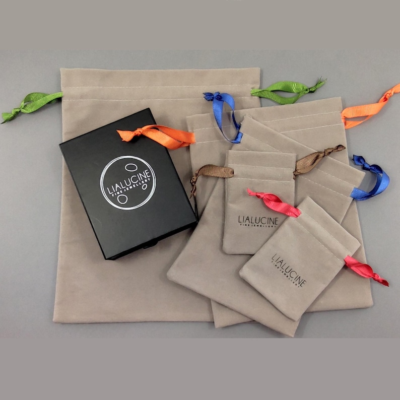 May include: An assortment of five beige velvet drawstring pouches with different coloured ribbons. The pouches are arranged in decreasing size, with the largest pouch at the back and the smallest pouch at the front. The pouches are all labelled with the text "LIALUCINE FINE JEWELLERY".