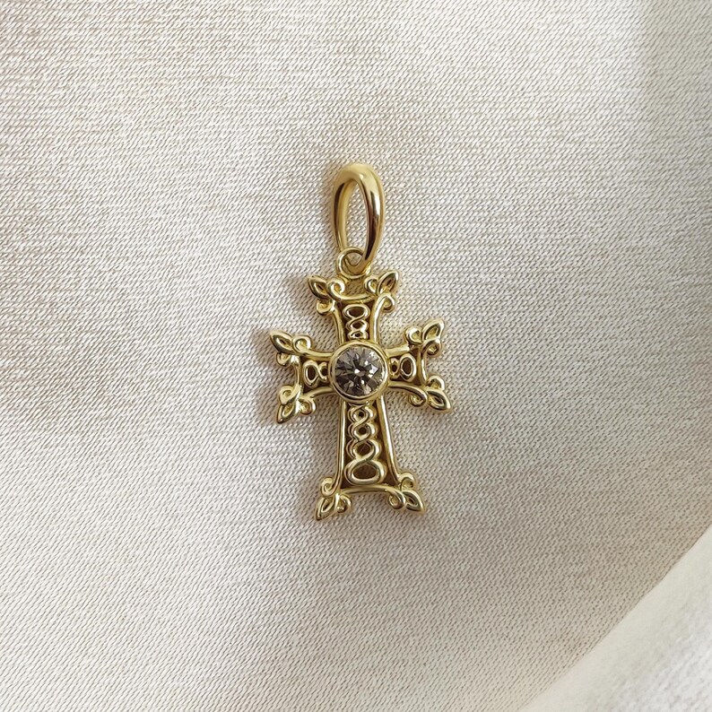 May include: A gold-toned cross pendant with a clear gemstone in the centre. The cross has intricate details and a looped design.