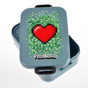 Op de afbeelding: Een blauwe lunchbox met een gepixelde hartvorm en de tekst "YOUR NAME" op de voorkant. De lunchbox is open en de binnenkant is zichtbaar.