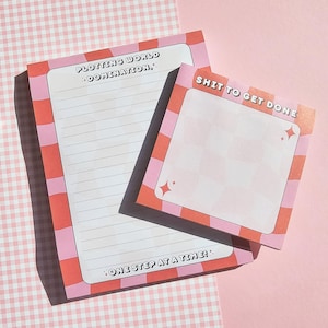 Op de afbeelding: Twee notitieblokken met roze en rode geblokte randen. Eén notitieblok heeft gelijnd papier en de tekst "Plotting World Domination, One Step at a Time!". De andere heeft een geblokt patroon en de tekst "Shit to Get Done".