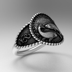 May include: A silver ring with a detailed design featuring two fish intertwined. The ring has a textured band and a decorative border.