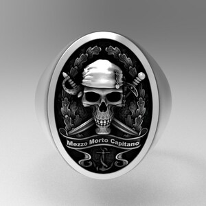 Peut inclure: Une bague en argent avec un crâne noir portant un bandana et tenant des épées croisées. Le crâne est entouré d'une couronne de feuilles et les mots "Mezzo Morto Capitano" sont inscrits sur une bannière sous le crâne. Une ancre se trouve sous la bannière.