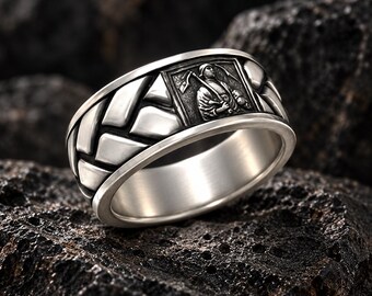 May include: A silver-toned metal ring featuring a wide band with a raised, braided pattern and a rectangular inset panel. The panel displays a detailed relief engraving of a figure holding a scythe over their shoulder, finished with dark oxidation.