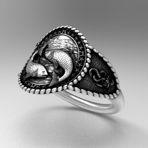 May include: A silver ring with a detailed design of two fish swimming in a circle. The ring has a rope-like border and a textured surface.
