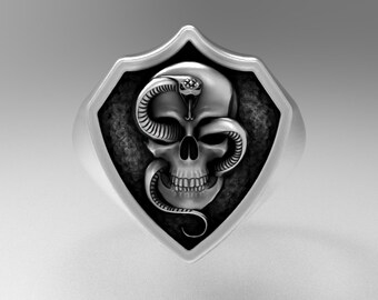 May include: A silver ring with a shield-shaped design featuring a skull with a snake wrapped around it.