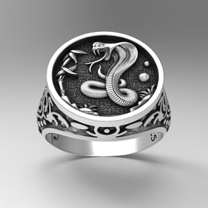 Könnte beinhalten: Ein silberner Ring mit einem detaillierten Design einer Kobra-Schlange mit einer erhabenen, strukturierten Oberfläche. Die Schlange ist um einen Stab gewickelt, der mit einer Blume gekrönt ist. Der Ring hat eine strukturierte Band mit einem floralen Design.