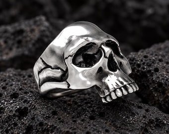 May include: A silver-toned ring shaped like a human skull. The ring features a polished, reflective surface with detailed eye sockets, a nasal cavity, and a row of teeth. The band of the ring is integrated into the sides of the skull design.