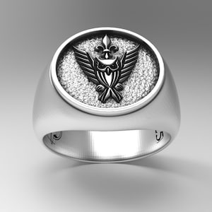 May include: A silver ring with a round, textured design featuring a black owl with a fleur-de-lis on its head. The ring has a smooth band.