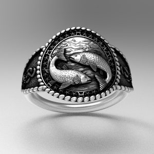 May include: A silver ring with a detailed design of two fish swimming in a circle. The ring has a textured band and a black background.