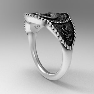 May include: A silver ring with a detailed design featuring a snake, an anchor, and a bird. The ring has a textured band and a raised design.
