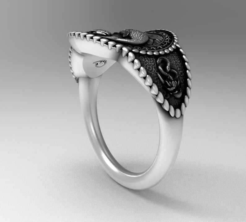May include: A silver ring with a detailed design featuring a snake, an anchor, and a bird. The ring has a textured band and a raised design.