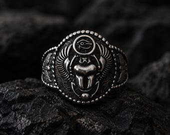 May include: A silver-toned ring featuring a central scarab beetle motif with outstretched wings. Above the beetle is an Eye of Horus symbol. The ring has a beaded border and a textured, dark oxidised finish, showcasing intricate metalwork details.