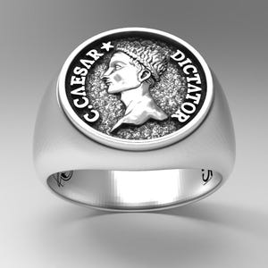 May include: A silver ring with a round, raised design featuring a profile of a person's head with the text "C.CAESAR*DICTATOR" around the edge.
