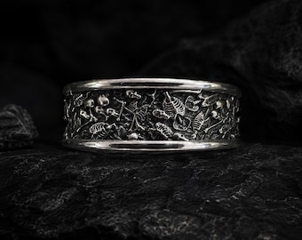 May include: A wide silver band ring featuring a detailed, textured relief pattern of multiple human skulls and skeletal rib cages across the entire outer surface, finished with polished, smooth edges along the top and bottom of the band.