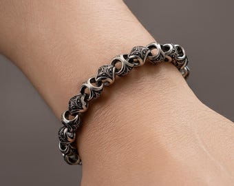 May include: Silver bracelet with an ornate, repeating pattern of interlocking links. The bracelet is made of a shiny, metallic material and is worn on a wrist. The design features detailed, symmetrical shapes, creating a classic, elegant look.