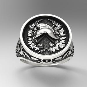 May include: A silver ring with a detailed design. The ring features a central emblem with a helmet, crossed axes, and laurel leaves. The band has an ornate, swirling pattern.