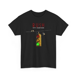 Op de afbeelding: Zwart T-shirt met de bandnaam "RUSH" in rood boven een verkeerslichtafbeelding. Het verkeerslicht toont rode, gele en groene lichten. De woorden "FIFTY SOMETHING" staan onder de bandnaam. Twee vogels zitten op een draad.