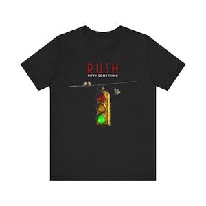 以下が含まれることがあります： 黒いTシャツ。赤い文字で「RUSH」と「FIFTY SOMETHING」と書かれています。デザインは、緑色のライトが点灯している信号機、電線上の鳥、そして飛んでいる鳥です。