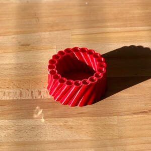 May include: A red 3D printed cylindrical container with a circular opening at the top. The container is made of many small, vertical tubes that are arranged in a spiral pattern.