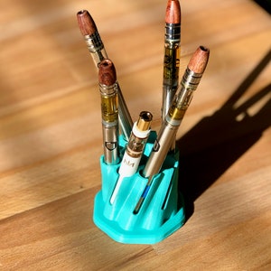 May include: A teal blue 3D printed stand with five slots for holding vape pens. The stand has a unique, wavy design. The vape pens have wooden tips and clear glass cartridges.