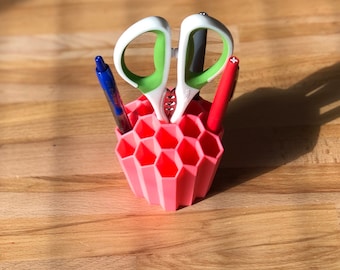 Op de afbeelding: Een roze zeshoekige bureau-organizer met een paar groene en witte scharen, een blauwe pen en een rode pen.