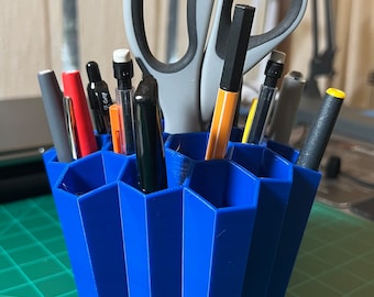 Op de afbeelding: Een blauwe zeshoekige bureau-organizer met meerdere vakken bevat verschillende schrijfbenodigdheden, waaronder pennen, potloden en een schaar.