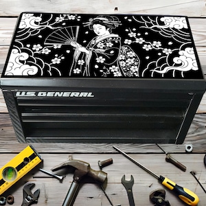 Può includere: Cassetta degli attrezzi nera con un piano decorativo con un design di geisha in bianco e nero. La cassetta ha il testo "U.S. GENERAL" sul davanti. Vari strumenti, tra cui una livella, un martello e chiavi inglesi, sono disposti intorno alla cassetta.