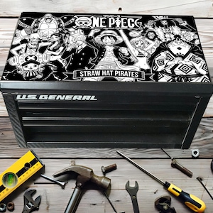 Peut inclure: Coffre à outils noir US General avec un graphisme noir et blanc One Piece Straw Hat Pirates sur le couvercle. Divers outils, dont un niveau, un marteau et une clé, sont disposés autour du coffre sur une surface en bois.